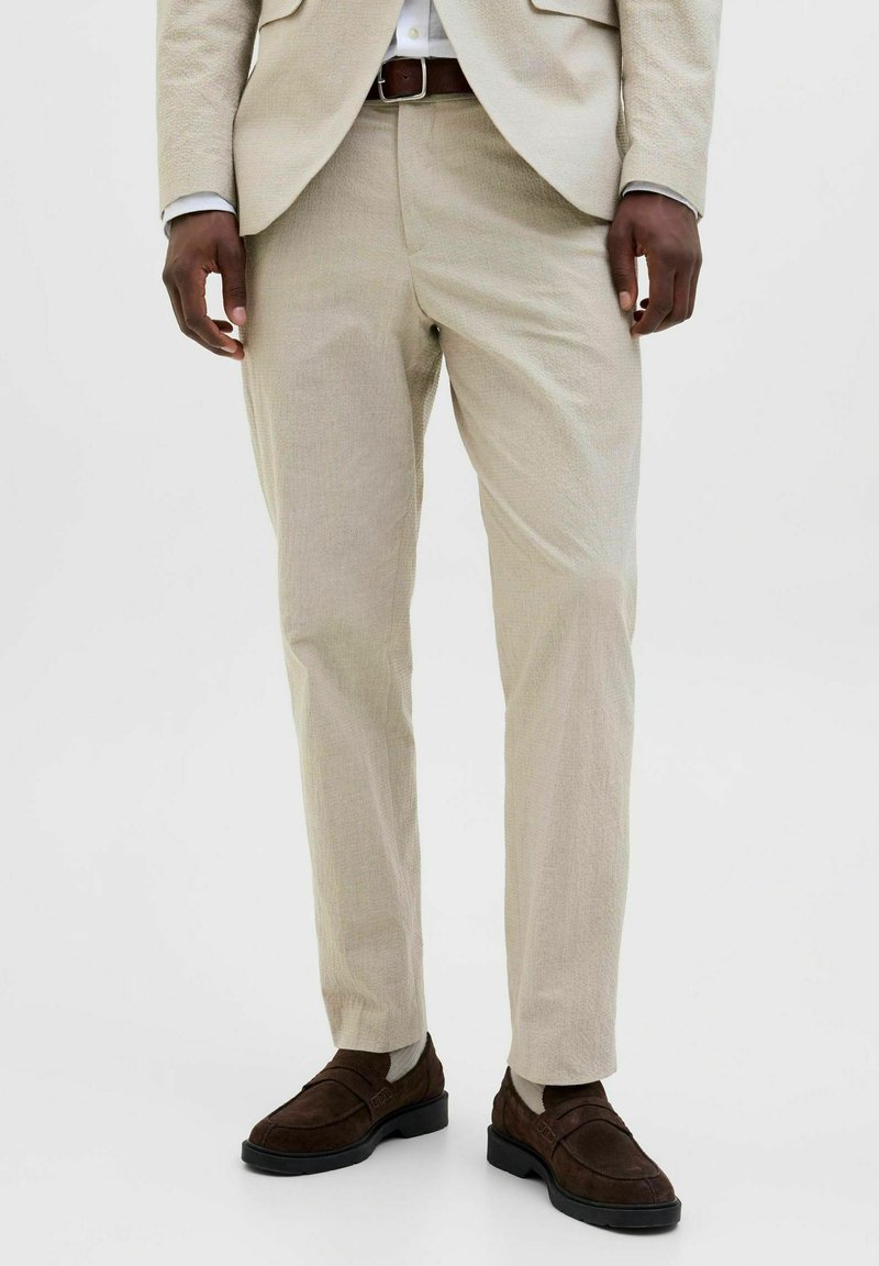 Lichtbeige op maat gemaakte broek van textuurstof, slim fit, in combinatie met donkerbruine nubucklederen loafers en een bruine riem. Heeft zichtbare zakken en naden.