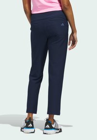 Μπλε σκούροι cropped αθλητικοί παντελόνες με λείο ύφασμα, διαθέτουν πίσω τσέπες και διακριτικό λογότυπο στη δεξιά πλευρά, σε συνδυασμό με μαύρα και λευκά παπούτσια.