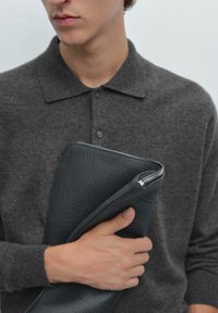 Schwarzer Leder-Dokumentenhalter mit strukturierter Oberfläche, teilweise geöffnet, gehalten gegen ein dunkelgraues Woll-Poloshirt mit Kragen und Knöpfen.