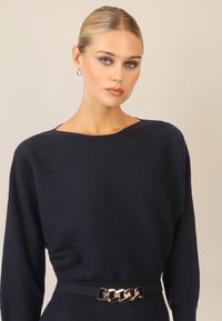 Mørkeblå strikket sweater med bådudskæring, lange ærmer og en struktureret overflade; har et bredt stofbælte med guldkæde hardware.