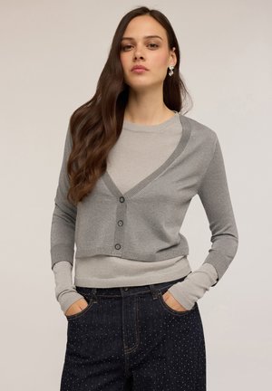 Hellgrauer, taillierter Cardigan mit V-Ausschnitt, Knopfverschluss und langen Ärmeln; kombiniert mit dunkelblauen Jeans, die kleine weiße Punkte aufweisen.