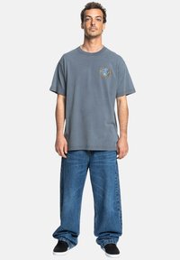 Gray oversized cotton t-shirt with a colorful circular graphic on the left chest, paired with wide-leg blue denim jeans and black shoes.
