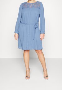 Modré šaty s dlouhým rukávem, zdobené krajkou kolem výstřihu, s vypasovaným pasem a splývavou sukní. Doplněné stříbrnými sandály na řemínky.