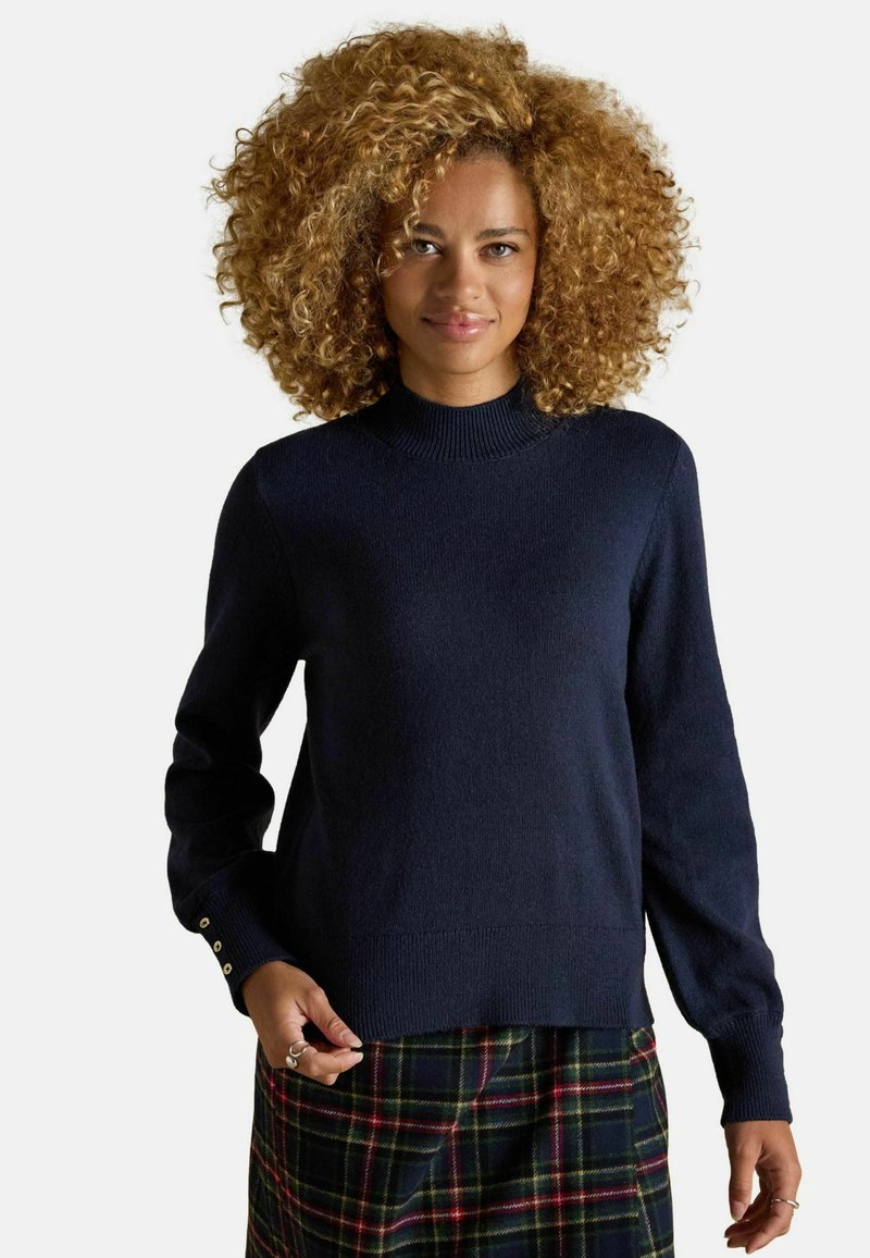 Marineblauer, gestrickter Pullover mit hohem Ausschnitt, gerippten Bündchen und vier Akzenten aus goldenen Knöpfen an den Ärmeln, kombiniert mit einem karierten Rock.