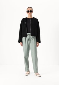 Giacca cropped nera con texture e zip argentata, abbinata a pantaloni a vita alta verde chiaro e scarpe beige senza lacci. Occhiali da sole neri indossati.