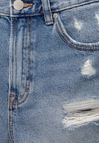 Siniset kulutetut farkut, joissa on rispaantuneita kohtia ja valkoisia lankoja. Kangas on haalistettu ja siinä on oransseilla saumoilla korostettu tikkauksia.