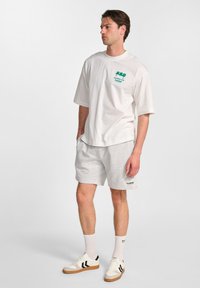 Witte katoenen t-shirt met groene grafische tekst, gecombineerd met lichtgrijze shorts. Inclusief witte sokken en beige sneakers met zwarte strepen.