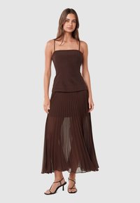 Jupe maxi plissée marron avec superposition transparente associée à un haut ajusté sans manches marron. Présente des bretelles spaghetti et une texture lisse.