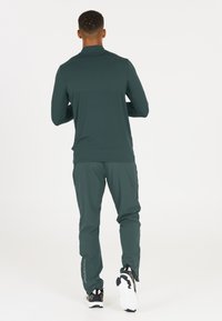 Mörk teal långärmad topp kombinerad med matchande avsmalnande byxor. Plagget har en slät textur och en atletisk passform, vilket gör det lämpligt för aktiv träning.