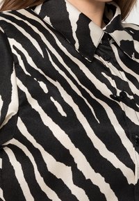 Blusa con stampa zebra nera e crema, caratterizzata da una texture satinata liscia, colletto a punta e bottoni neri lungo la chiusura frontale.