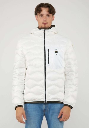 Jonge man met kort haar die een witte doorgestikte jas met zwarte rits en borstzak draagt, gecombineerd met blauwe spijkerbroek, staand met het gezicht naar voren.