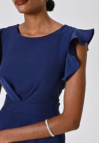 Marineblaue Kleid mit Rüschensäulen, präsentiert ein V-förmiges Taillendetail, glattes Material, kombiniert mit einem glänzenden silbernen Armband.
