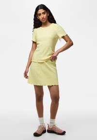 Femme aux longs cheveux bouclés portant un ensemble jaune composé d'un haut à manches courtes et d'une jupe, avec des baskets roses et des chaussettes blanches sur fond blanc.