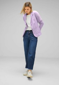Lavender blazer with two front pockets, worn over a white top and paired with wide-leg blue jeans and light sneakers, standing with hands clasped.
