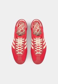 Zapatillas deportivas rojas con un upper texturizado, que presentan un diseño de tres rayas en color crema, cordones planos y un collar acolchado.