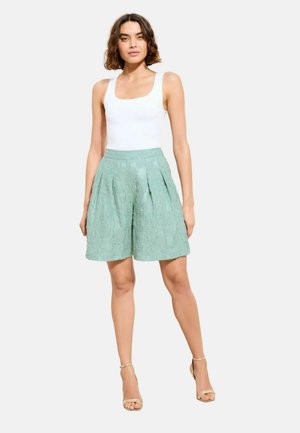 Kvinde står iført en hvid ærmeløs top, mintgrøn tekstureret shorts og beige højhælede sandaler på en ensfarvet baggrund.