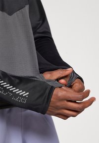 Długorękawowa koszulka sportowa w gradientowym odcieniu szarości i czerni. Posiada otwory na kciuki i teksturowaną tkaninę z nadrukowanym logo na mankiecie.