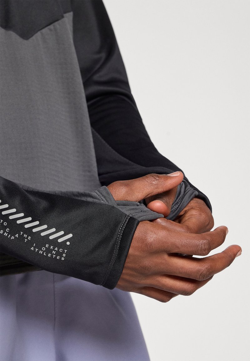 Langærmet atletisk top i gradient grå og sort. Har hul til tommelfingrene og tekstureret stof med trykt branding langs ærmet.