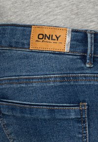 Jeans de denim azul con una etiqueta de cuero que presenta "ONLY" y "Blue Denim anno 1995". Incluye costuras visibles y un acento plateado.