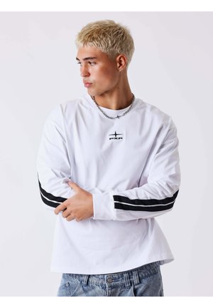 Jeune homme aux cheveux blonds courts regarde à gauche, portant une chemise blanche à manches longues avec des rayures noires sur les manches et un jean bleu.