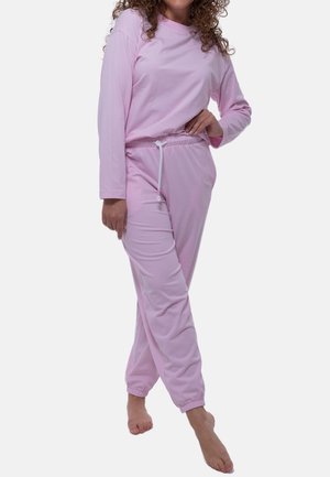 Woman in light pink long-sleeve top and matching jogger pants standing barefoot with one hand on her hip against a plain background.