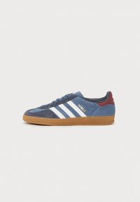 Sapatilhas de camurça azul com riscas brancas e texto dourado "Gazelle". Apresenta uma sola de borracha castanha, colarinho acolchoado e material texturizado.