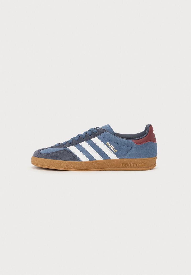Sapatilhas de camurça azul com riscas brancas e texto dourado "Gazelle". Apresenta uma sola de borracha castanha, colarinho acolchoado e material texturizado.