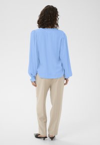 Blouse bleu clair à manches longues avec épaules bouffantes, encolure froncée, associée à un pantalon beige et des chaussures à talons noires.