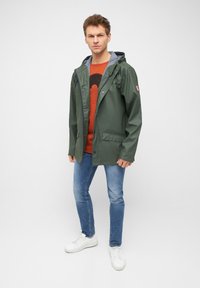 Giacca impermeabile verde con cappuccio, chiusura con zip e bottoni a pressione, due tasche frontali e fodera a righe. Indossata sopra una maglietta grafica arancione.