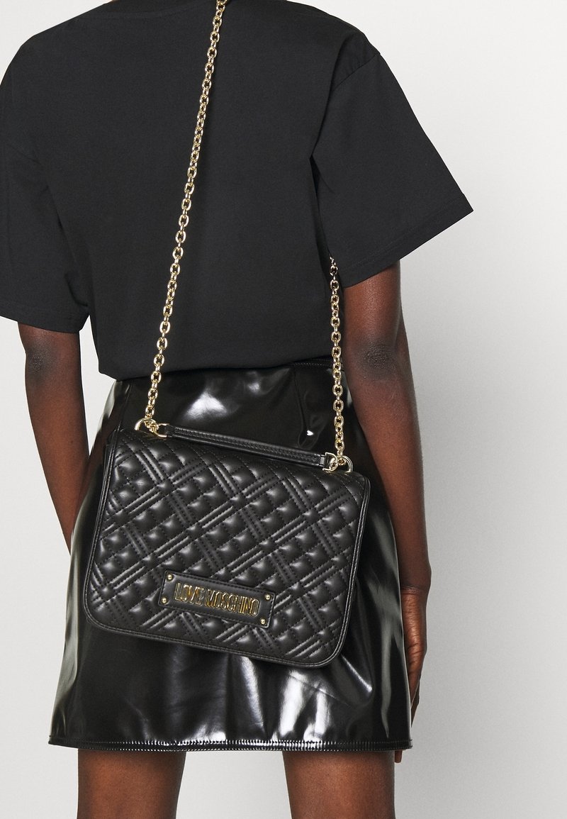 Černá prošívaná kožená kabelka přes rameno se zlatým řetízkem a emblémem "Love Moschino", nošená přes černý top s krátkým rukávem a lesklou černou sukni.