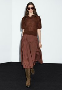 Maglione corto a maniche corte marrone, abbinato a una gonna fluida marrone e stivali al ginocchio in suede marrone. La modella indossa occhiali da sole oversize.