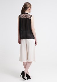 Blusa nera trasparente con schienale in pizzo, abbinata a culottes color crema. Modella che indossa tacchi alti neri, in piedi contro uno sfondo chiaro.