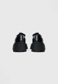 Black leather shoes with a chunky black rubber sole, featuring textured accents and elastic loops at the heel for easy wearing.