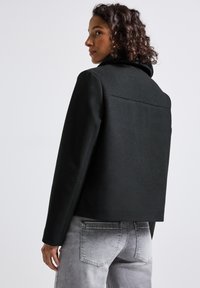 Donna con capelli ricci che indossa una giacca nera corta e jeans chiari lavati, che guarda di lato su uno sfondo neutro.