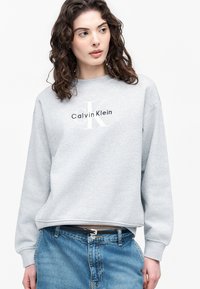 Femme aux cheveux bouclés portant un sweat-shirt Calvin Klein gris clair et un jean bleu, regardant légèrement vers sa gauche.