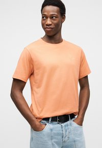 Κοντομάνικο μπλουζάκι από βαμβάκι σε ανοιχτό πορτοκαλί χρώμα με στρογγυλή λαιμόκοψη και διακριτική λεπτομέρεια λογότυπου. Συνδυασμένο με τζιν παντελόνι σε ανοιχτό μπλε και μαύρη ζώνη.