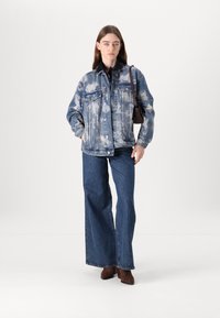 Denimjakke med skylignende mønstre, frontknapper og to brystlommer; brukt med vide denimjeans og brune ankelstøvler.