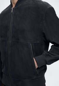Άνδρας που φοράει σκούρο μπλε bomber μπουφάν με ριμπ λαιμόκοψη και μανσέτες, φερμουάρ στο μπροστινό μέρος και χέρι στην πλαϊνή τσέπη με φερμουάρ.
