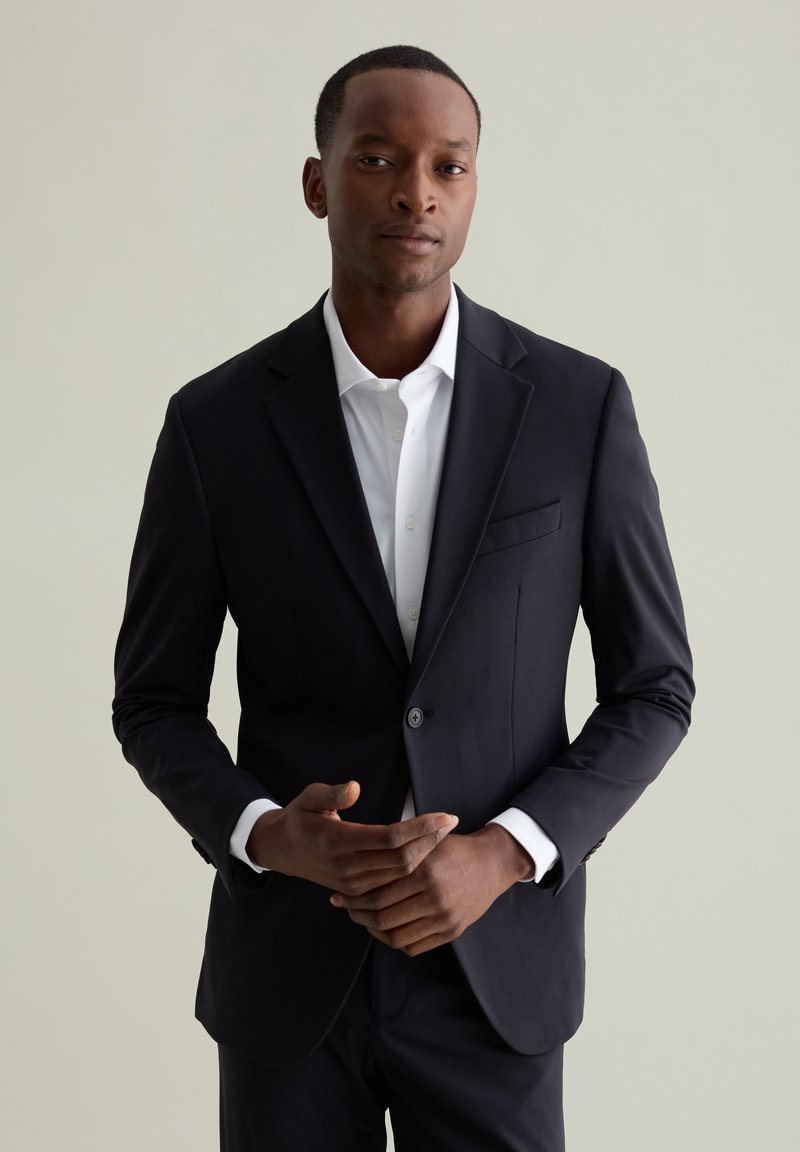 Blazer noir avec un bouton à l'avant, revers crantés et une poche poitrine. Porté par-dessus une chemise blanche, d'une coupe ajustée et d'une texture lisse.
