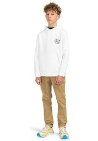 Vit hoodie med en färgglad grafik på bröstet, kombinerat med beige byxor och flerfärgade sneakers. Modellen står i en avslappnad pose.