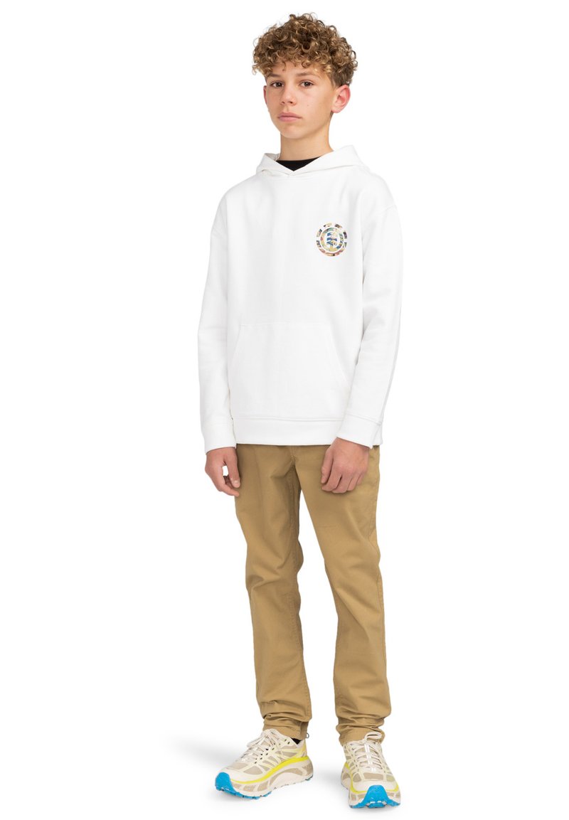 Vit hoodie med en färgglad grafik på bröstet, kombinerat med beige byxor och flerfärgade sneakers. Modellen står i en avslappnad pose.