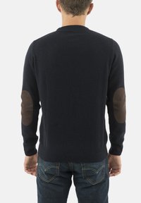 Mężczyzna w granatowym swetrze z brązowymi łatek na łokciach i ciemnoniebieskich dżinsach, pokazany od tyłu na białym tle.