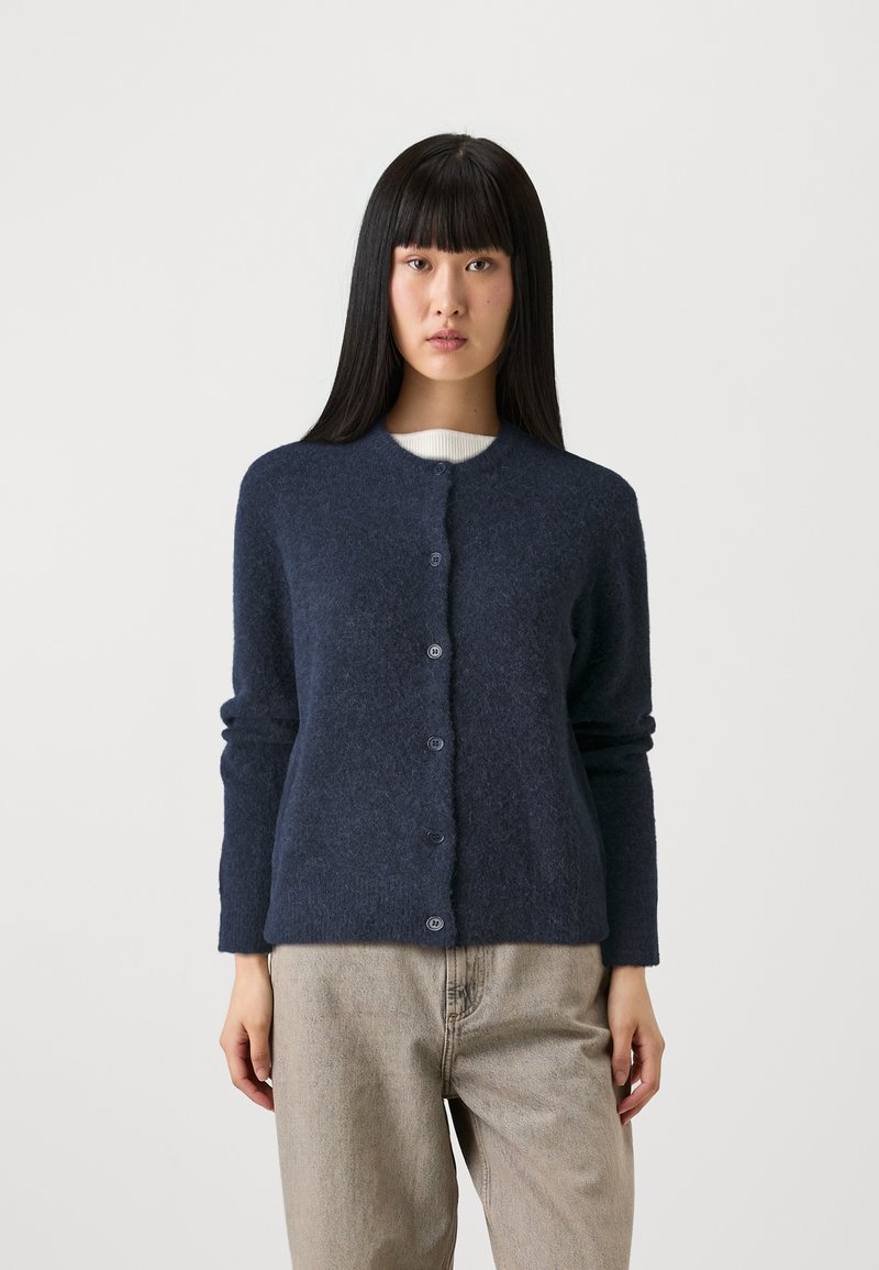 Woman with long black hair wearing a navy blue buttoned cardigan over a white top and light beige pants, standing against a plain white background.