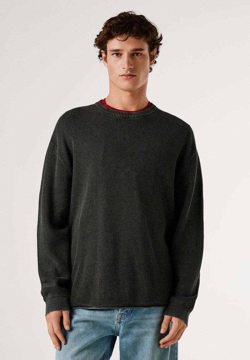 Pull en maille gris foncé avec un col rond et des manches longues, présentant un motif texturé. Porté sur un haut rouge.