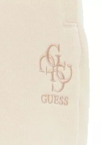 Beige sweatpants met een zachte textuur, voorzien van een geborduurd "GUESS" logo en een overlappend letterontwerp aan de linkerkant.