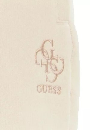 Pantaloni della tuta beige con una texture morbida, caratterizzati da un logo ricamato "GUESS" e un design di lettere sovrapposte sul lato sinistro.