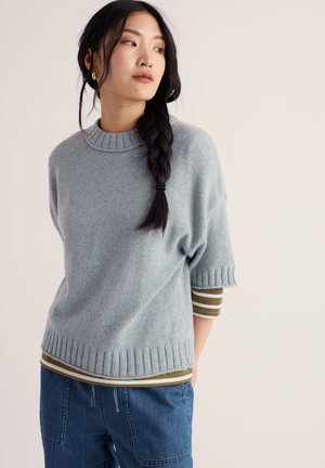Donna con lunghi capelli scuri intrecciati indossa un maglione chiaro grigio ampio, una camicia a maniche a righe e jeans blu, guardando di lato.