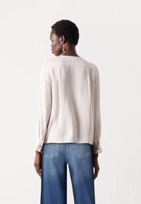 Lys beige langermet bluse med en jevn tekstur, som har sirkulær skjæring nederst og knapper på mansjettene, sammen med vide blå denimjeans.