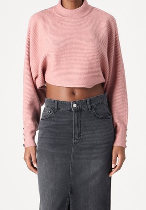 Vrouw draagt een roze geribbelde cropped trui met knoopdetails aan de mouwen en een donkergrijze denim rok met hoge taille en een split aan de voorkant.