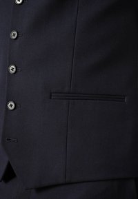Blazer bleu marine en tissu texturé, avec trois boutons et une poche plate. Le design présente une couture subtile et une coupe classique ajustée.